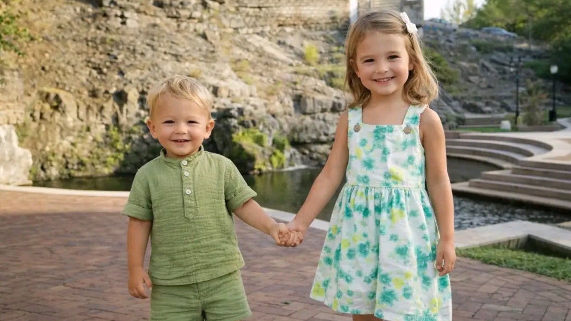 Two children, a boy and a girl, holding hands outdoors with a stone wall and water feature in the background.