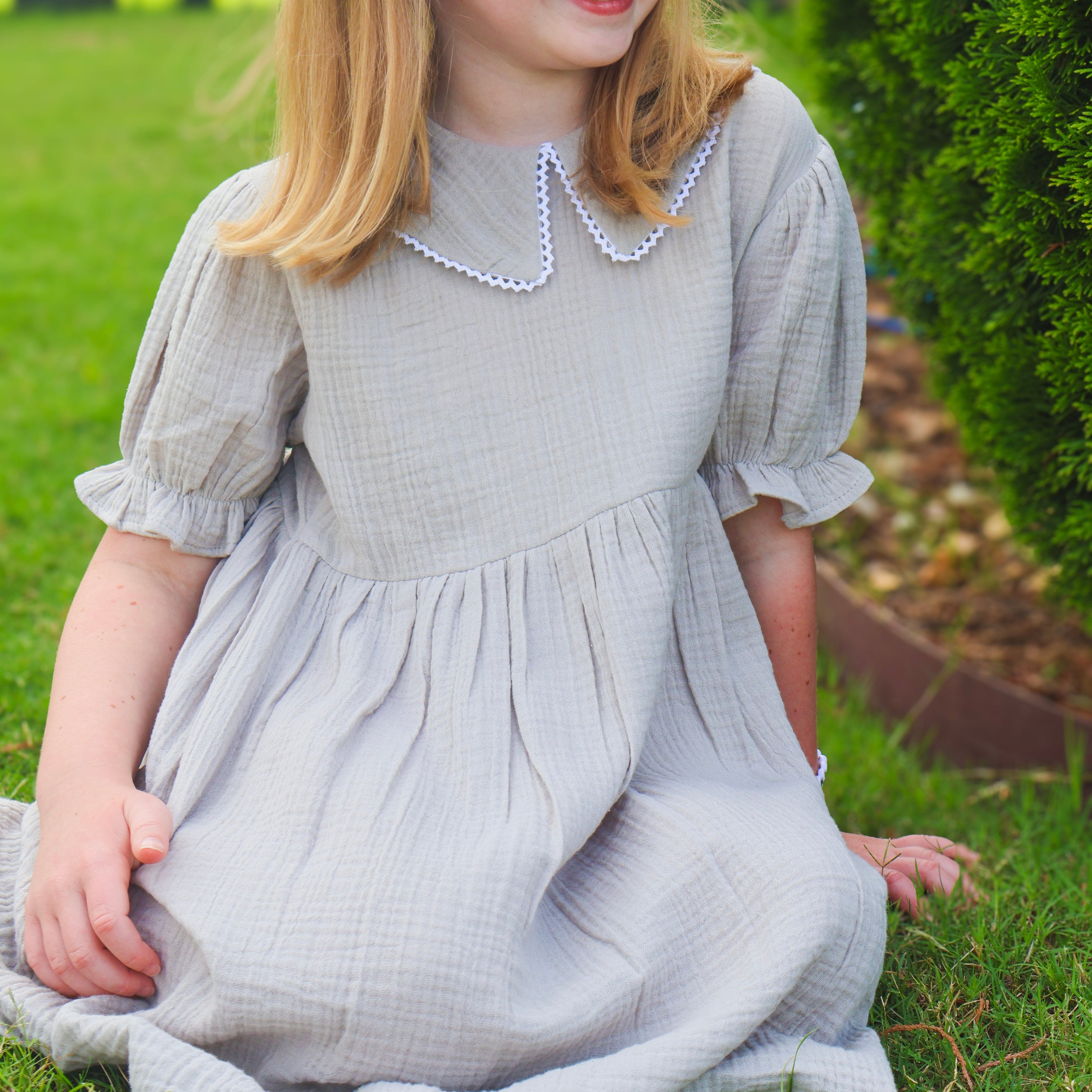 A young girl with blonde hair sits on a green lawn wearing a light gray textured cotton dress with a white beaded Peter Pan collar and a matching bow headband.