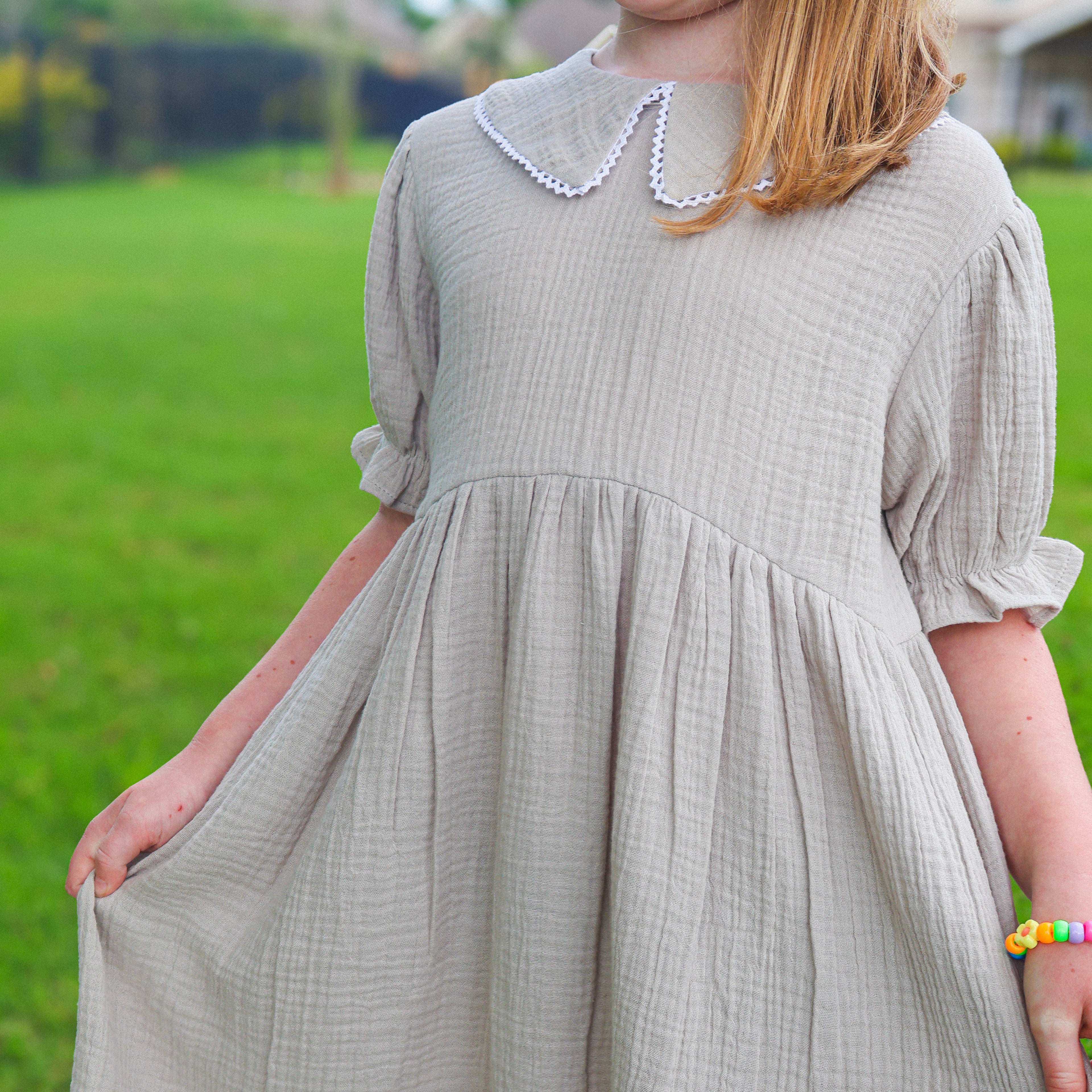 A young girl wearing a light gray textured cotton gauze dress with a white beaded Peter Pan collar, standing outdoors on a green grass lawn.