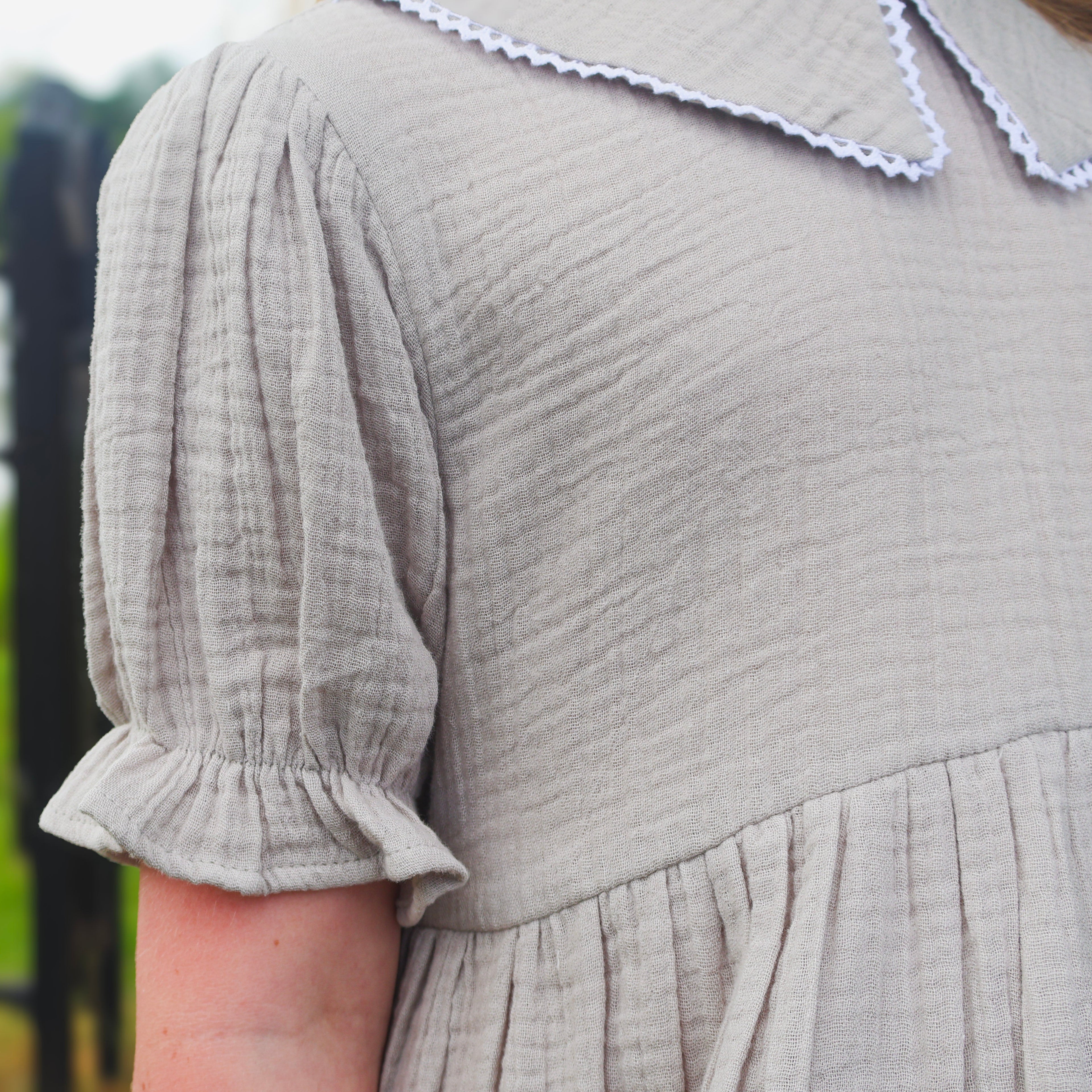 Close-up of a person wearing a light beige textured cotton dress with short puff sleeves, ruffled cuffs, and a white scalloped Peter Pan collar, standing outdoors in front of a blurred fence and greenery.